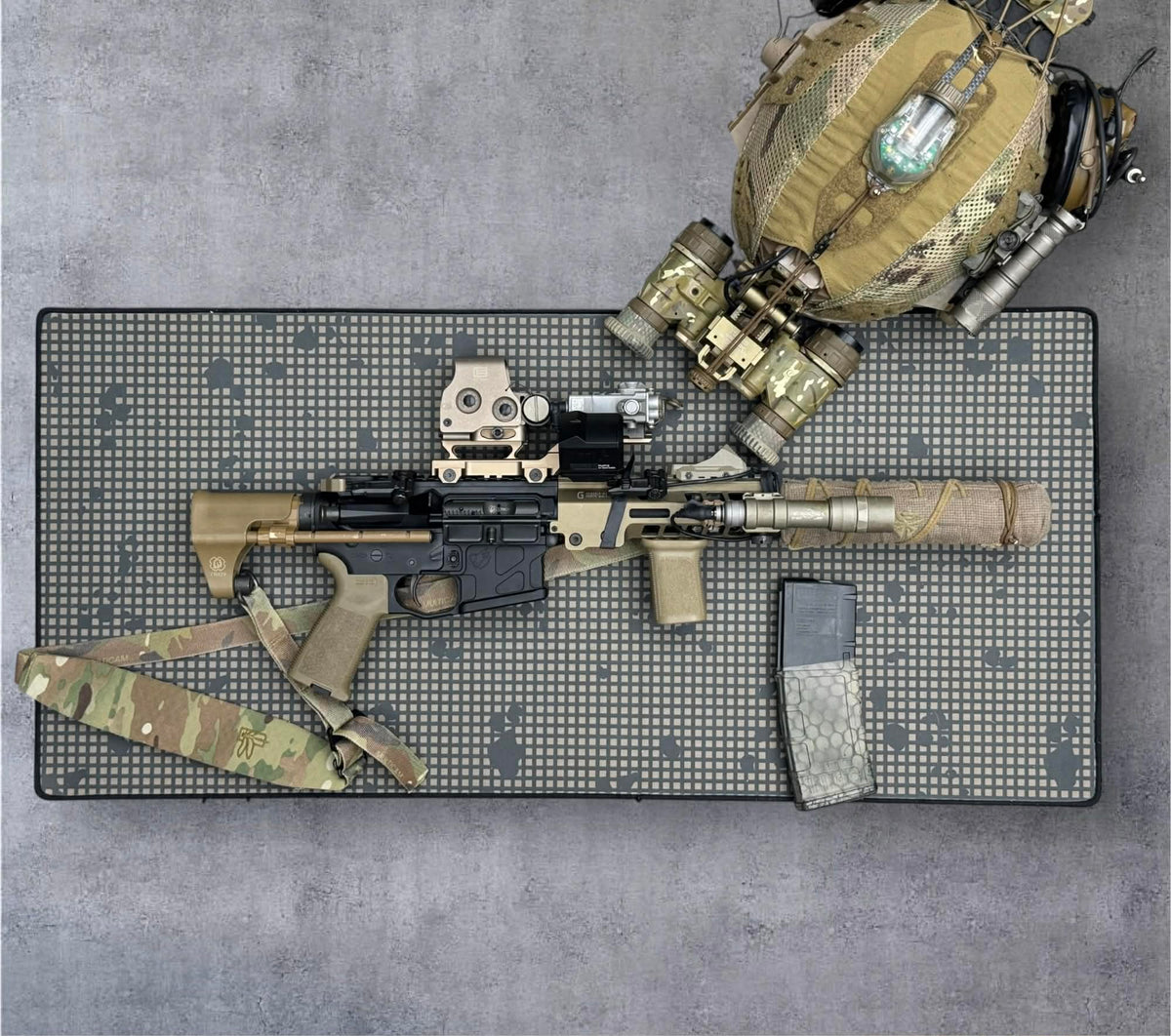 A tactical rifle laid flat on a gray mat with a black dotted border, alongside a matching camouflage sling, magazine, and a tan tactical backpack or gear bag on a concrete-like gray background.